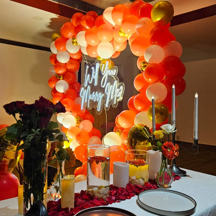 Romantic marriage proposal decoration in Hyderabad with a heart‑shaped balloon arch, glowing “Will You Marry Me” LED neon sign, and rose petal carpet.