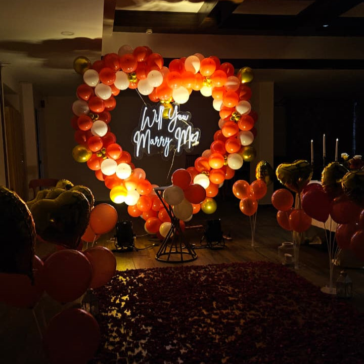 Romantic marriage proposal decoration in Hyderabad with a heart‑shaped balloon arch, glowing “Will You Marry Me” LED neon sign, and rose petal carpet.