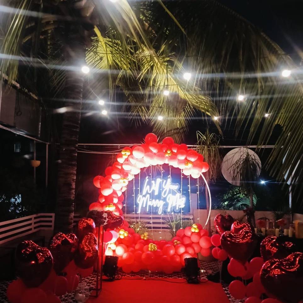 A romantic rooftop setup with red balloons, heart decor, and a neon sign reading "Will You Marry Me" under string lights.