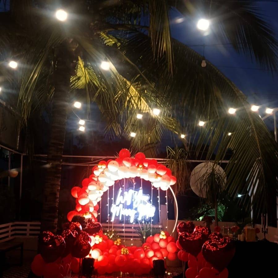 A romantic outdoor setup at night featuring a neon sign, red and white balloons, heart-shaped decorations, and string lights among palm trees.