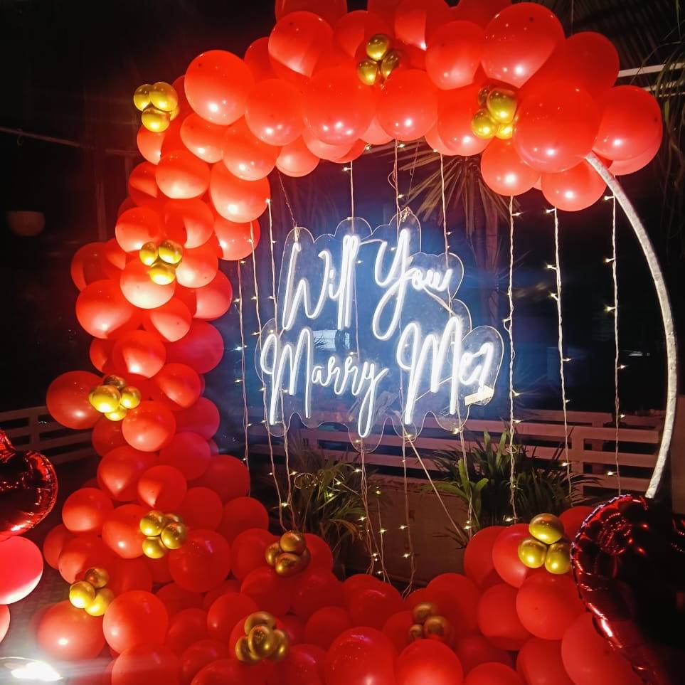 A romantic proposal setup featuring a neon sign reading "Will You Marry Me?" surrounded by vibrant red and gold balloons.