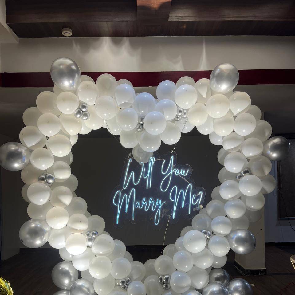 A heart-shaped balloon arch featuring white and silver balloons surrounds a glowing sign that reads "Will You Marry Me?"
