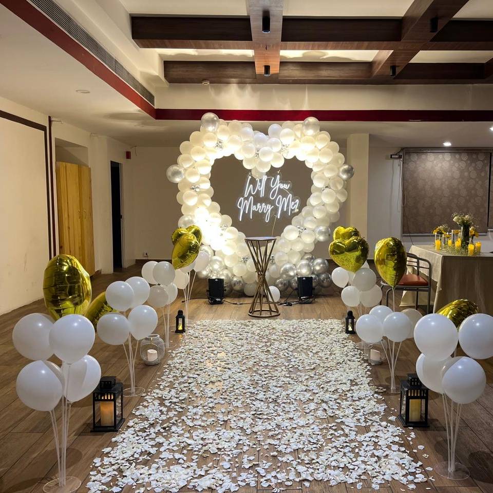 A romantic proposal setup featuring a heart-shaped balloon arch, white flower petals, and golden balloons, with a sign that reads "Will You Marry Me?"