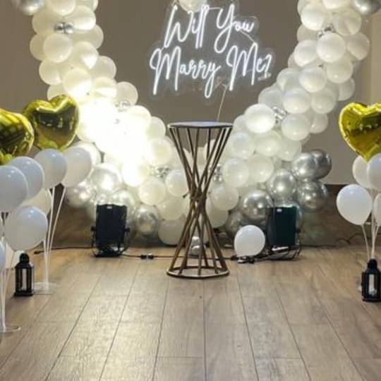 A romantic proposal setup featuring a neon "Will You Marry Me?" sign, white and silver balloons, and gold heart-shaped balloons.