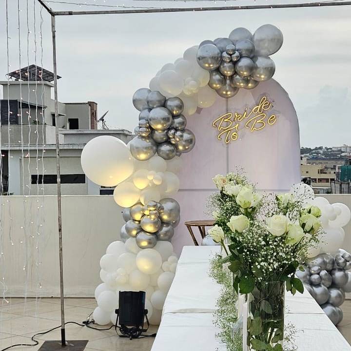A rooftop bridal shower setup with a "Bride To Be" sign, white and silver balloon arch, and a table decorated with white flowers.