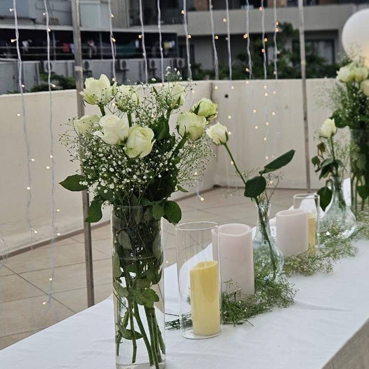 Elegant table setting for a bridal celebration, featuring white roses, green foliage, candles, and a "Bride to Be" neon sign with balloon arch.