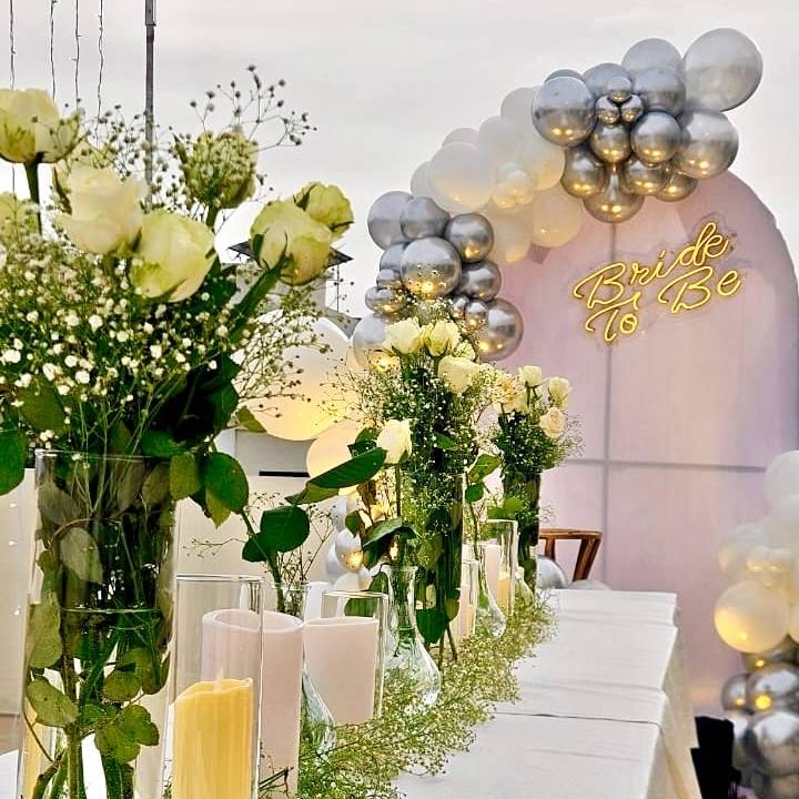 Elegant table setting for a bridal celebration, featuring white roses, green foliage, candles, and a "Bride to Be" neon sign with balloon arch.