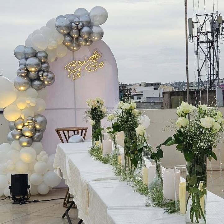 A stylish bridal shower setup featuring a balloon arch, white roses, candles, and a glowing sign that reads "Bride To Be."