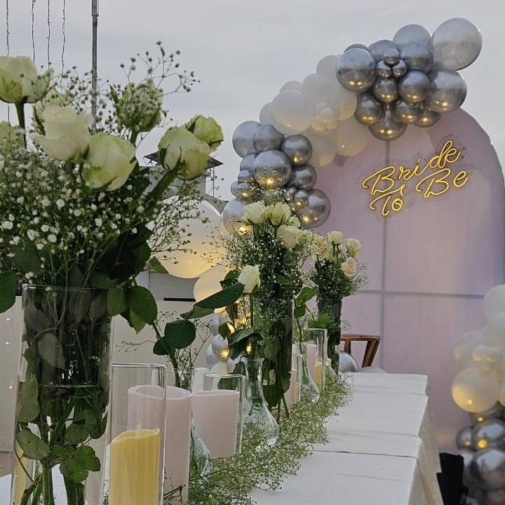 Elegant wedding setup featuring white roses, greenery, and candles, with a "Bride To Be" sign and balloon arch in the background.