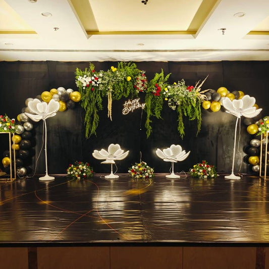 A festive birthday stage with a black backdrop, adorned with green foliage, flowers, and balloons in gold and black, featuring glowing floral displays.