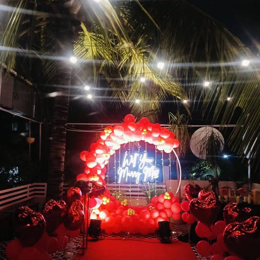 A romantic rooftop setup with red balloons, heart decor, and a neon sign reading "Will You Marry Me" under string lights.