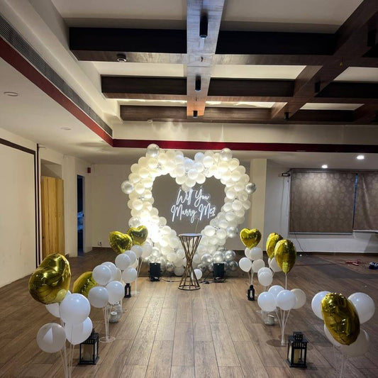 A romantic proposal setup featuring a heart-shaped arrangement of white and silver balloons, with "Will You Marry Me?" illuminated in the center.
