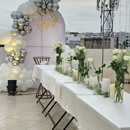 A rooftop setup with a long table decorated with white flowers and candles, and a "Bride To Be" sign surrounded by white and silver balloons in the background.