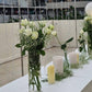 Elegant table setting for a bridal celebration, featuring white roses, green foliage, candles, and a "Bride to Be" neon sign with balloon arch.