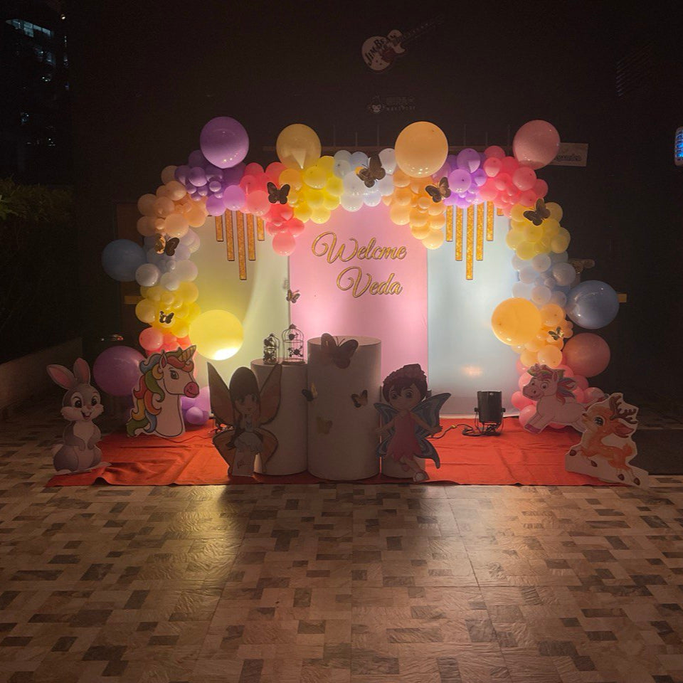 Pastel unicorn birthday party decoration with balloon arch, fairy cutouts, and customized welcome name backdrop setup